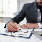 Geschäftsmann im Anzug analysiert Diagramme auf Papier und macht Notizen – Fokus auf Hand, Stift und Unterlagen mit Statistiken am Schreibtisch.