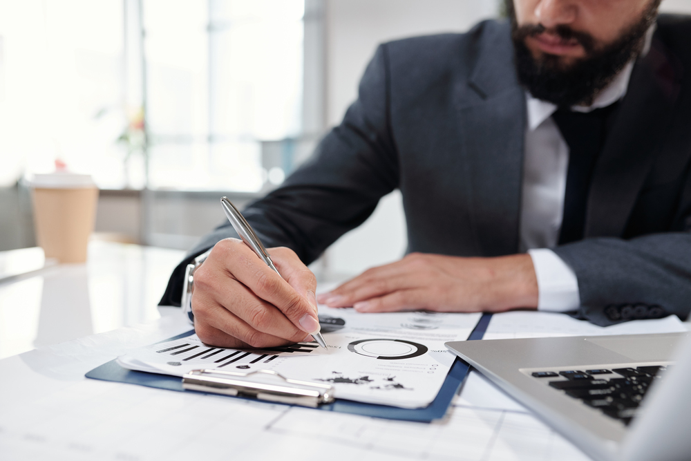 Geschäftsmann im Anzug analysiert Diagramme auf Papier und macht Notizen – Fokus auf Hand, Stift und Unterlagen mit Statistiken am Schreibtisch.