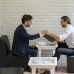 Zwei Männer in Business-Kleidung sitzen sich in einem modernen Café gegenüber und geben sich zur Begrüßung oder Einigung einen Fistbump – Symbol für partnerschaftliche Zusammenarbeit oder erfolgreiche Verhandlung.