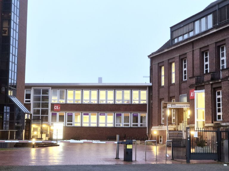 Eingangsbereich eines Backstein-Gebäudekomplexes im SMS Businesspark City bei Dämmerung, mit beleuchteten Fenstern und den Gebäudebezeichnungen A1 und C5.1.